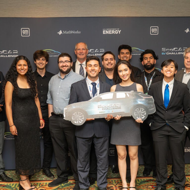 EcoCAR EV Challenge team group photo in front of event backdrop, with members smiling and holding a car-shaped sign.