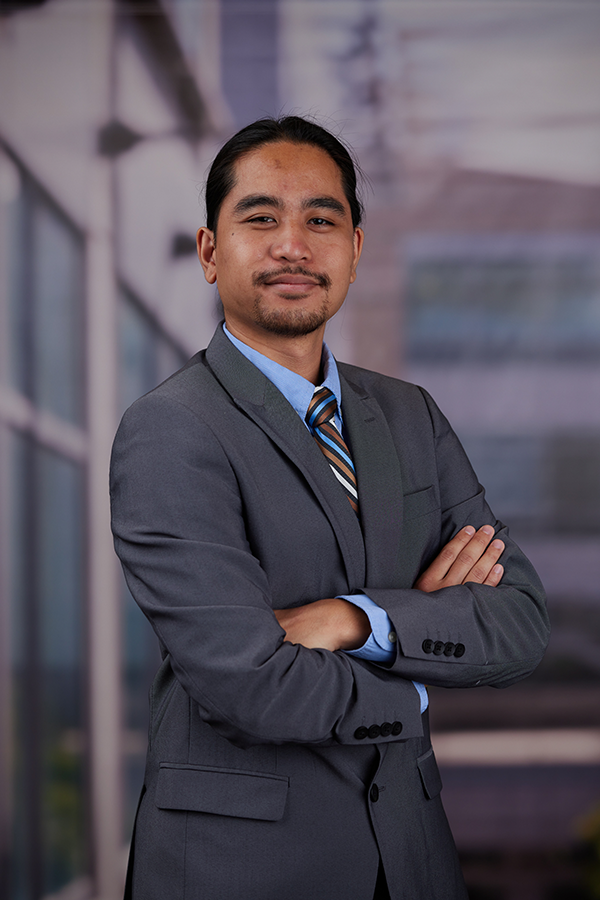 Male student wearing a grey suit with a blue shirt and striped tie with arms crossed looking straight into the camera. 