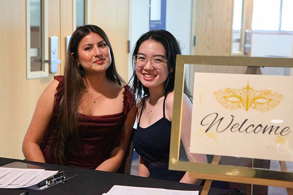 2025 Masquerade Gala two female students dressed in semi formal attire next to a check-in sign