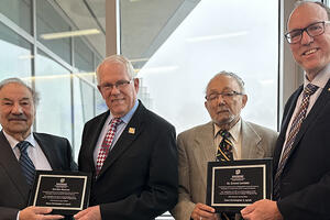 Four men wearing suits and ties are standing side by side, looking at the camera and smiling. From left to right:  Reza Abbaschian and Gordon Bourns hold a plaque, followed by Ernest Levister and Chris Lynch, also holding a plaque. The background features large windows with an outdoor view of trees and what appears to be a building in the distance.