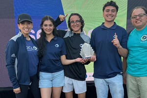 A diverse group of four students and MESA director standing together on a platform outdoors, holding a trophy and gesturing excitedly.