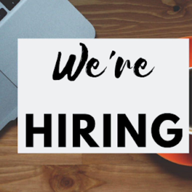 Text reads "We're Hiring" over an aerial view of a desk with a laptop and coffee