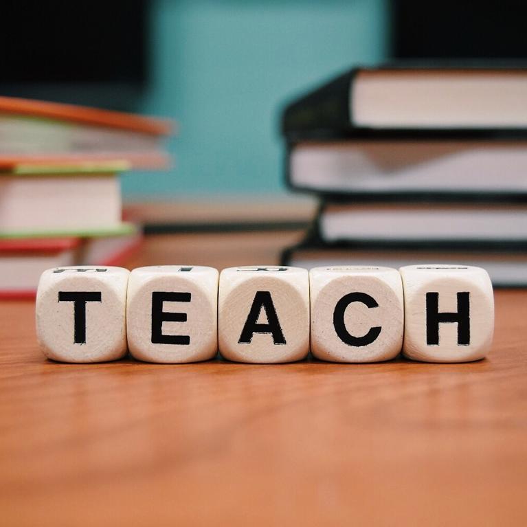 Four dice are aligned to spell the word "TEACH" on a desk with blurred books in the background.