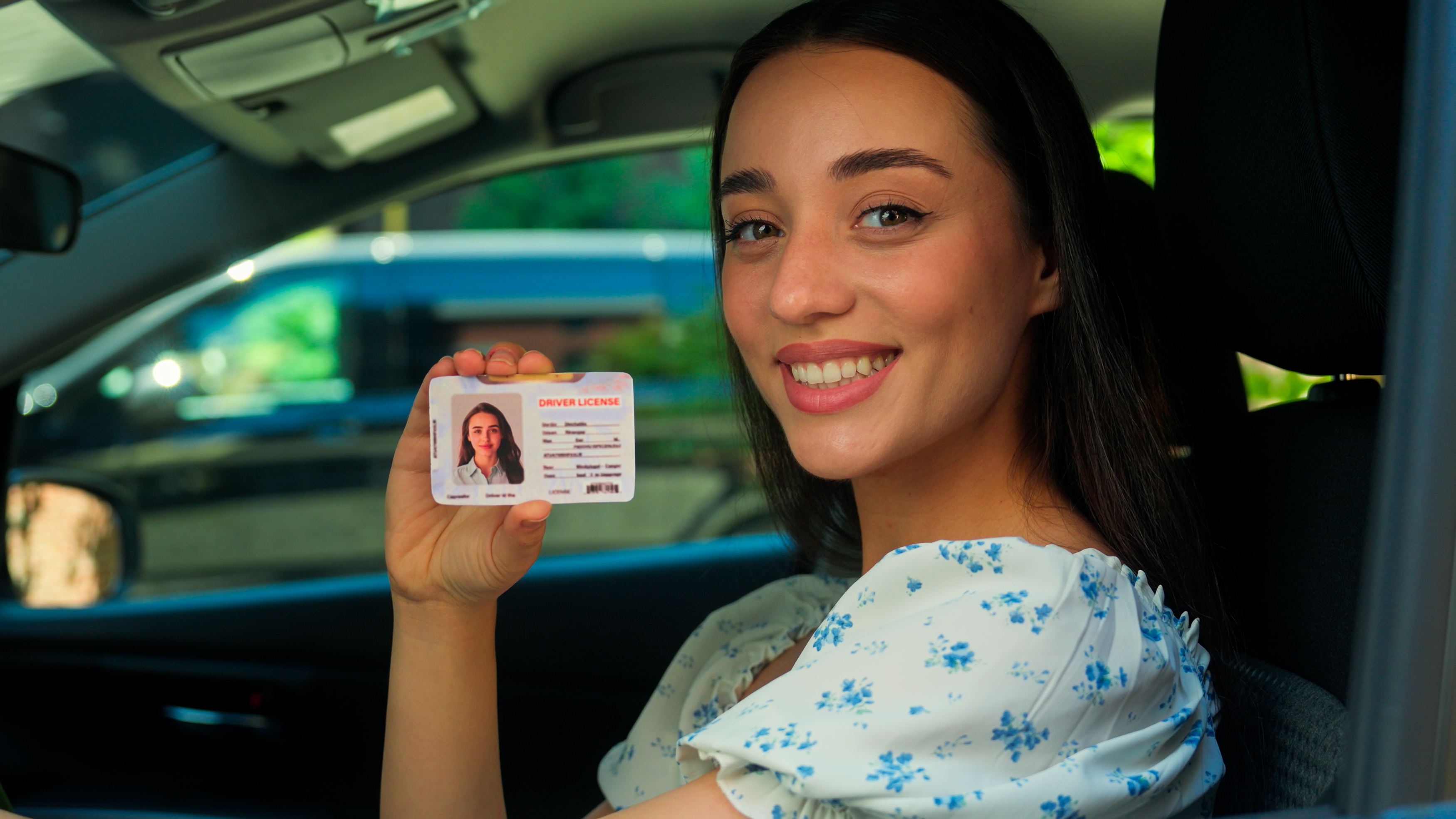 A young woman with long hair is smiling while holding up her driver's license as she looks out of her car window.