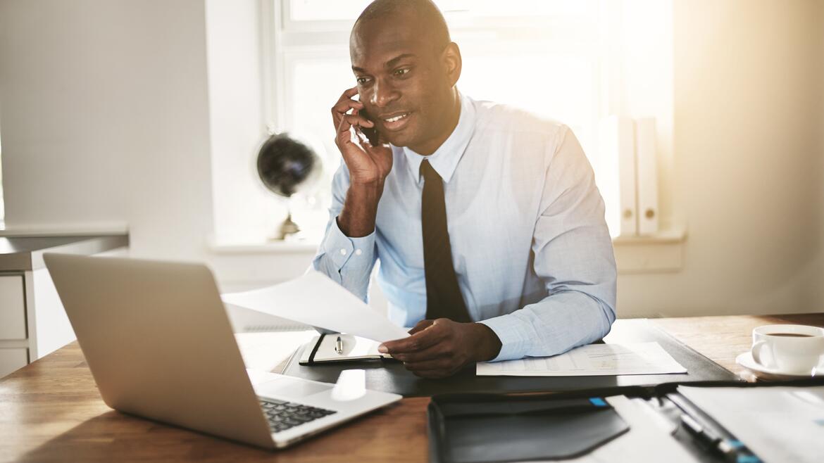 African American man talking on the phone, holding a sheet of paper with his left hand, and looking at a computer screen 