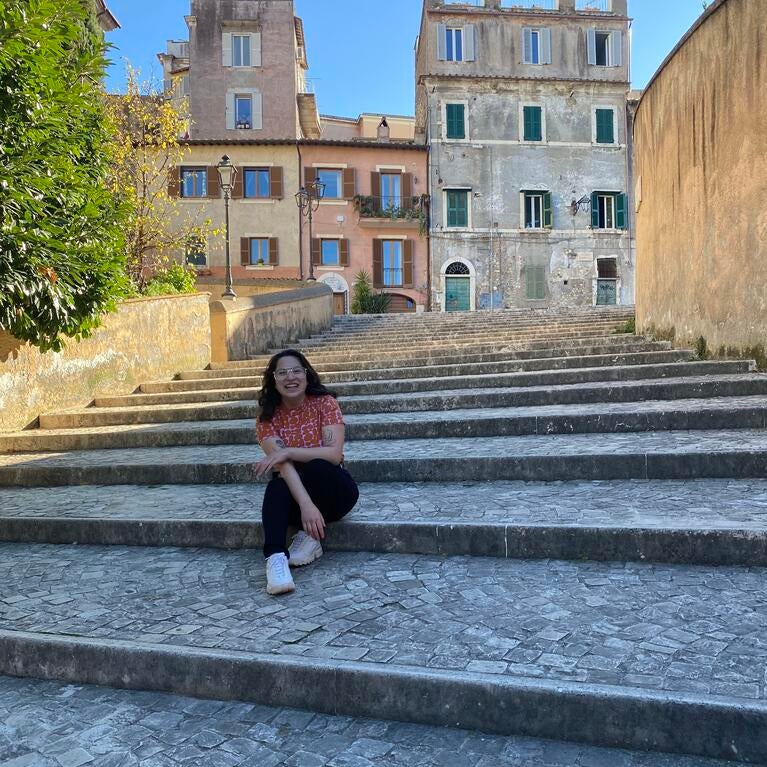 Female, early 20s, with dark hair, sitting on stone steps in front of old stone buildings in Italy