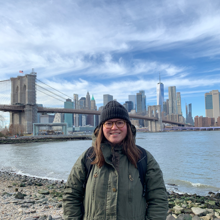 Female, early 20s, with brown hair, wearing a dark green winter coat, a black knitted winter hat, and glasses, standing near water, with the Brooklyn Bridge and the New York City Skyline behind