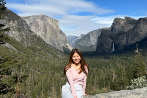 Female, early 20s, with dark hair, sitting on stone ledge in front of trees and mountain at Yosemite National Park