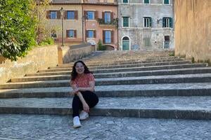 Female, early 20s, with dark hair, sitting on stone steps in front of old stone buildings in Italy