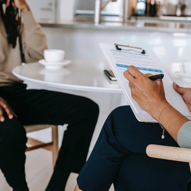 person fills out clipboard at table looking at other sitting person