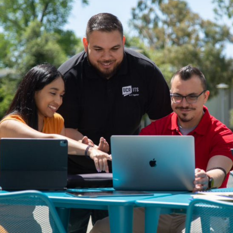 A worker in a black ITS Staff Shirt assisting a man and a woman with their laptops