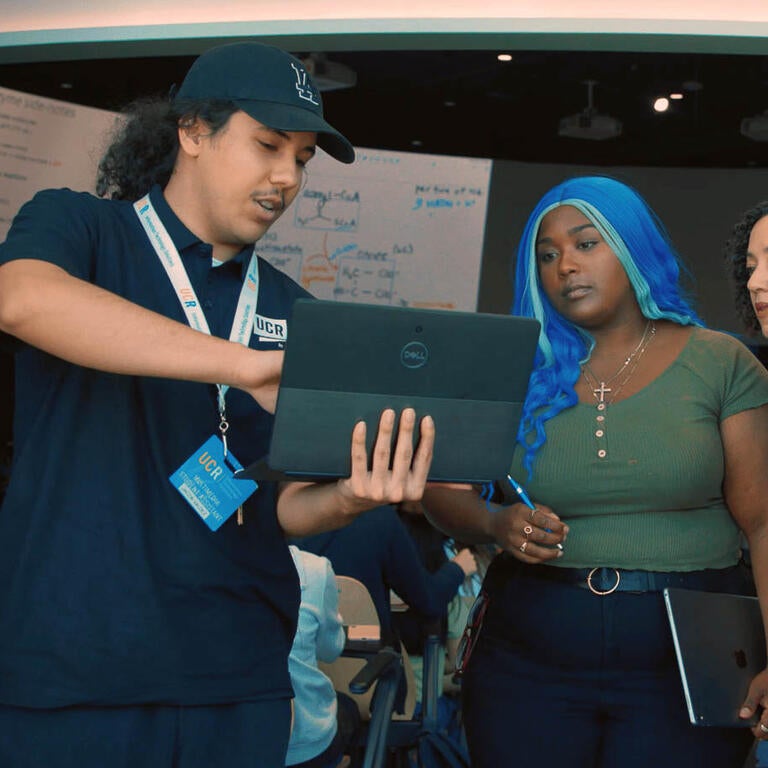 A student worker in a black shirt holding a laptop and showing the screen to four students