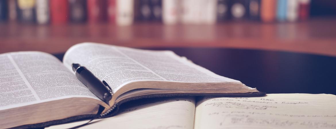 Two books open on table with books on a shelf in the background