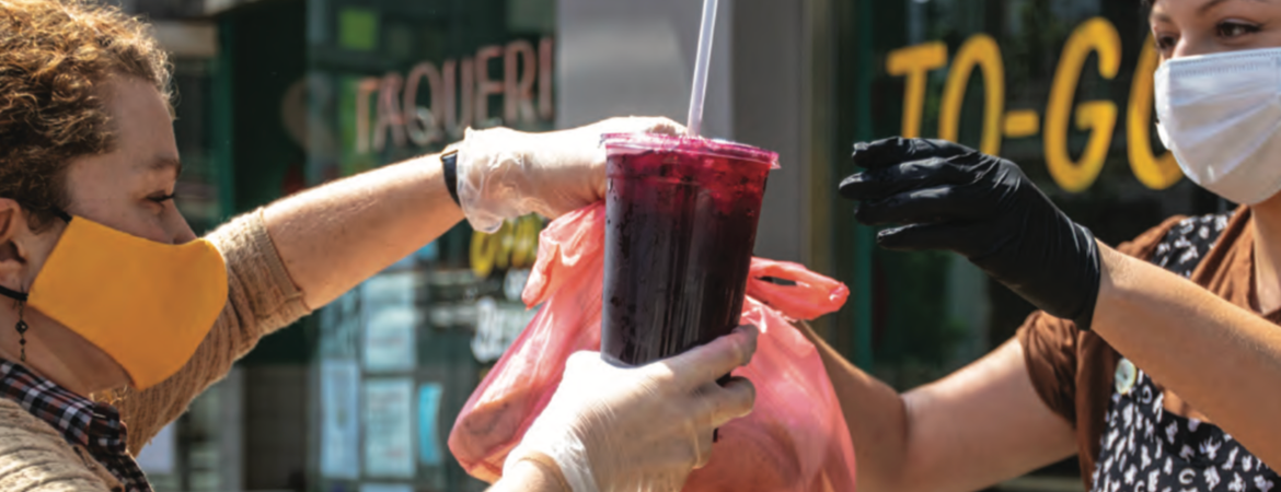 A shopkeeper hands a customer a drink while both wear masks and stay 6 feet apart