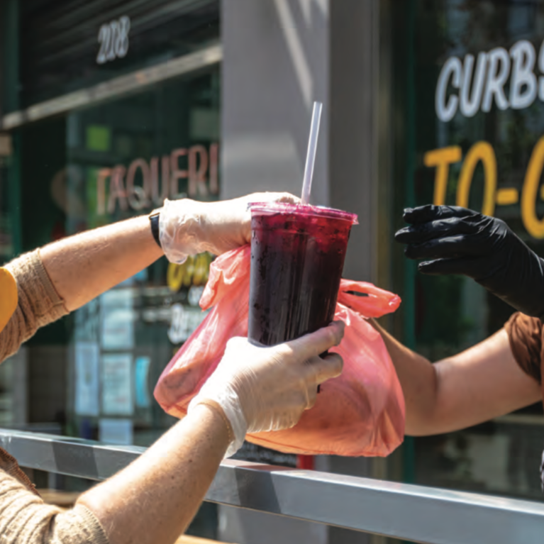A shopkeeper hands a customer a drink while both wear masks and stay 6 feet apart