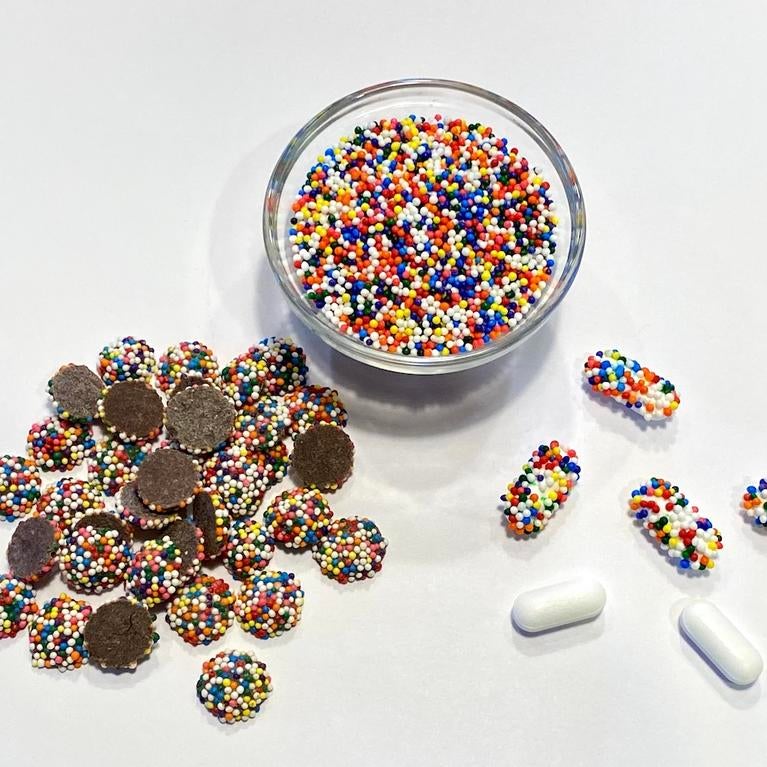 Chocolate drops covered with colorful candy nonpareils alongside pharmaceutical capsules also coated with the candy