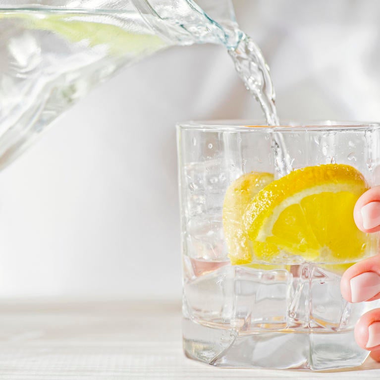 A person pours water from a pitcher into a glass with a lemon slice in it