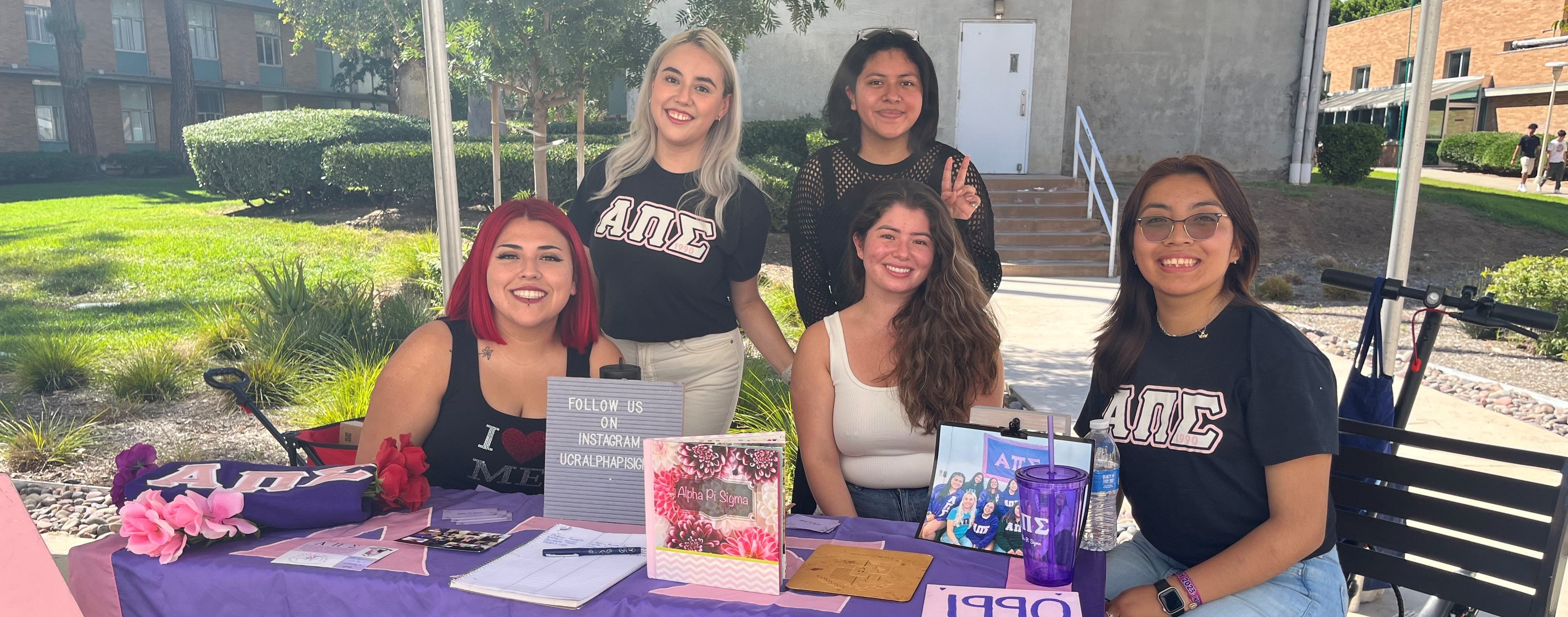5 members of Alpha Pi Sigma sitting behind a table that is decorated with their organization's letters