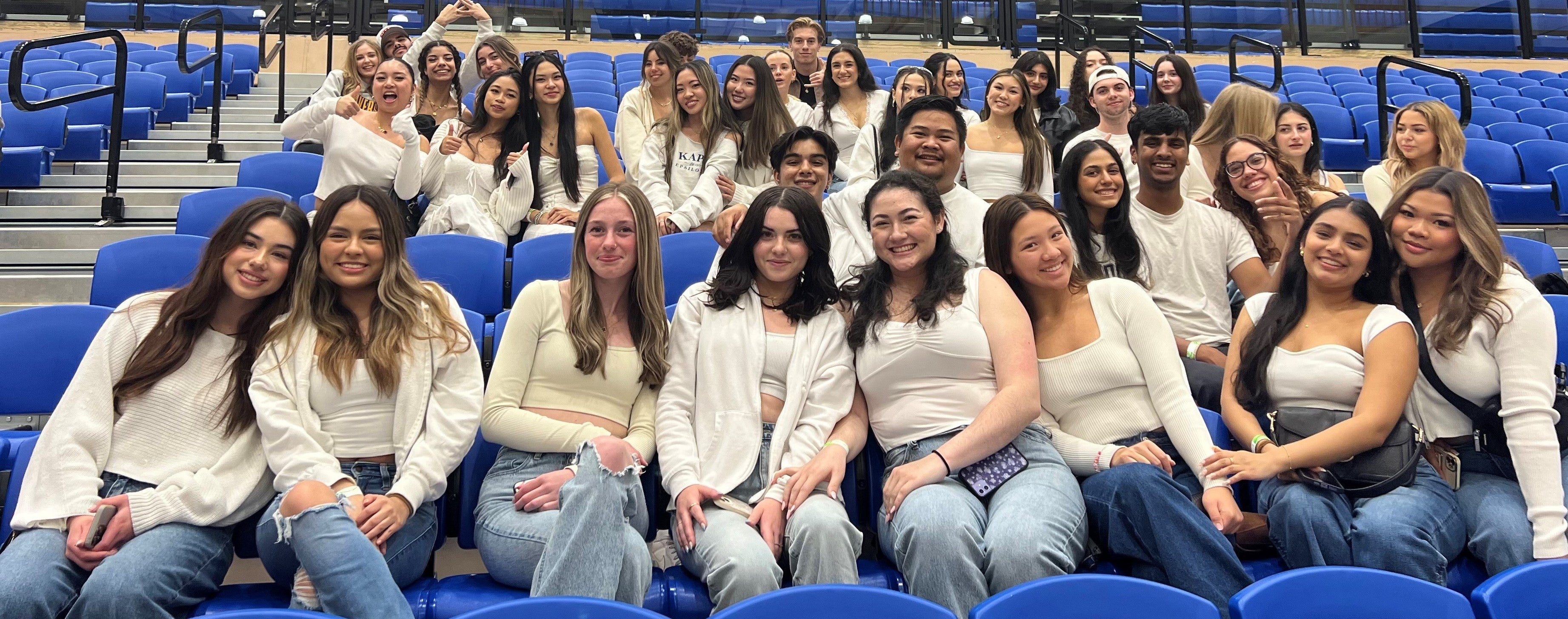 Rows of students sit in the MAC Gym stands, wearing white