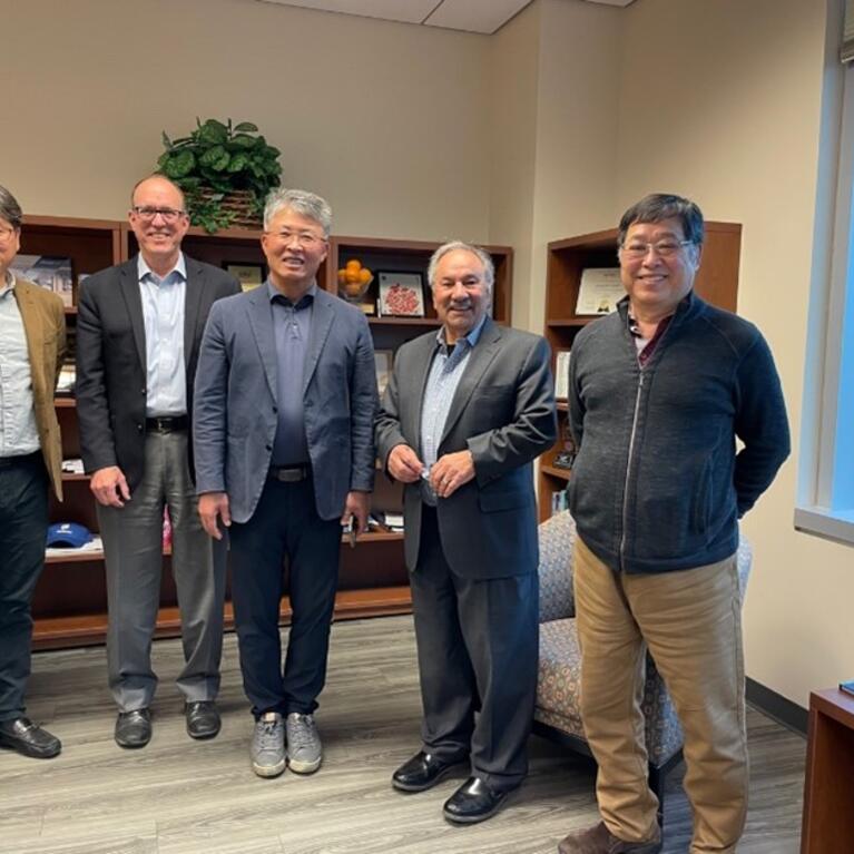  Five men are standing together in an office, smiling at the camera. They are dressed in business casual attire, and behind them is a bookshelf with various items, including plants and books.