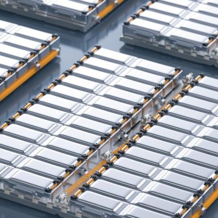  An aerial view showing an array of prismatic battery cells or modules arranged in rows on a clean, grey surface, suggestive of a dry room battery production environment. The silver-cased cells have a ribbed top surface and a narrow yellow band visible on the sides, indicating a high-volume manufacturing setting.