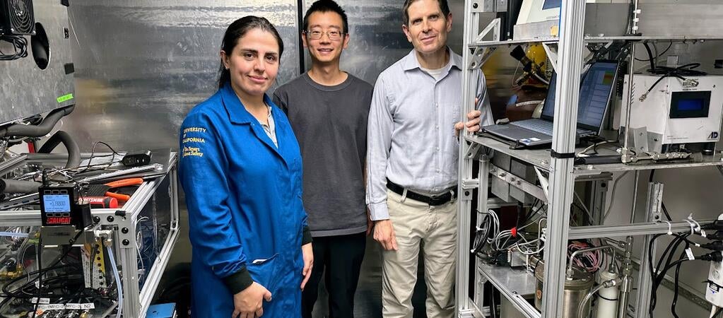 A front-view, full-body shot of three UCR CE-CERT researchers standing inside a mobile lab truck. From left to right: graduate student Maryam Yousefi, in a blue university jacket; graduate student Minghao Han, in a gray t-shirt and glasses; and CE-CERT Director Dr. Don Collins, in a collared shirt. They are all looking at the camera and smiling faintly. The mobile lab has shiny, corrugated metal walls and is filled with intricate scientific equipment on metal racks, with many wires visible.