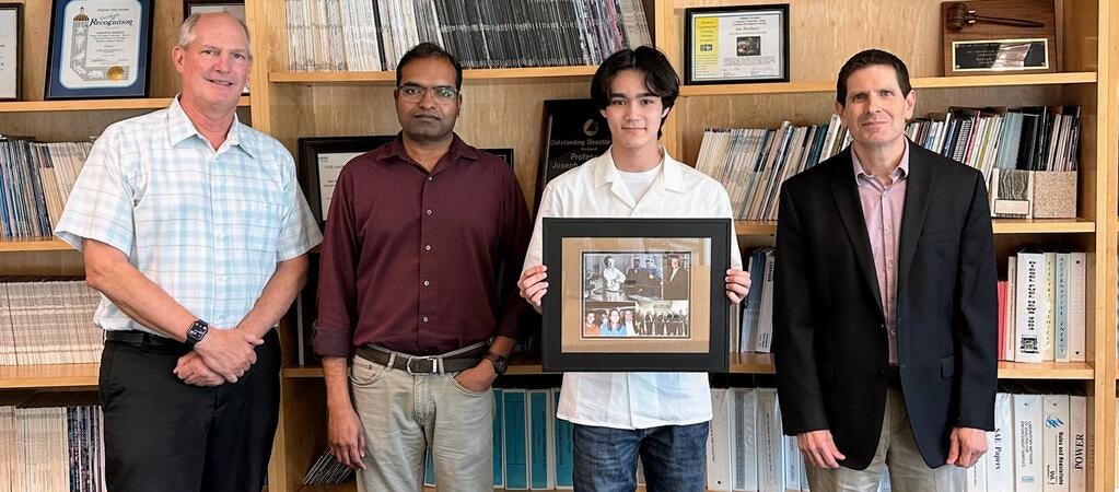 Warren Norbeck, a high school intern, stands holding a framed photo tribute to his grandfather, CE-CERT founder Dr. Joe Norbeck. He is flanked by three CE-CERT staff members: Arun Raju on his left, and Matt Barth and Don Collins on his right. All four are standing indoors in front of bookshelves filled with binders and reports.