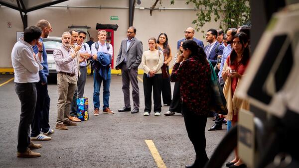A group of ZEV Working Group members gathers in a semicircle during a technical site tour of electric mobility infrastructure in Mexico City, with a speaker addressing the group while others listen attentively.