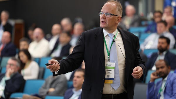 Professor Thomas Koch, keynote speaker from Karlsruhe Institute of Technology and CEO of Allianz Wasserstoffmotor, speaks to attendees during the Inaugural 2025 Hydrogen Engine Conference. He is holding a presentation clicker, wearing a suit and conference badge, with a full audience seated in the background.
