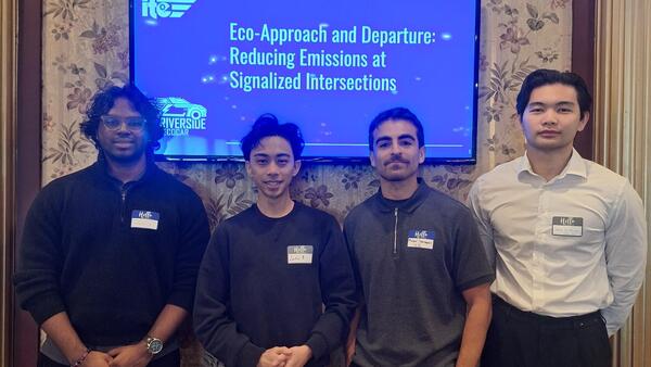 Four students standing in front of a presentation screen displaying the title 'Eco-Approach and Departure: Reducing Emissions at Signalized Intersections' at a UCR event. All are wearing name tags and business casual attire.