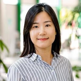 A woman with straight black hair, wearing a white button-up shirt with thin black stripes, smiles softly while standing in a bright indoor setting with plants in the background.