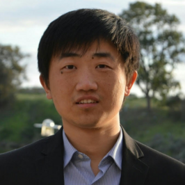 man wearing pinstriped collared shirt with black blazer looking at camera in front of park background