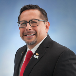 Alt text: A man wearing glasses, a black suit, white shirt, and red tie smiles at the camera against a blue background. He has short dark hair and a neatly trimmed goatee, with lapel pins visible on his jacket.