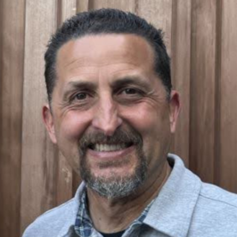 Man with grey beard and short spikey hair smiling in front of brown fence background wearing collared shirt