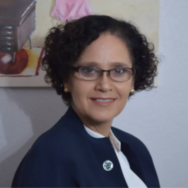 Woman with short curly hair with glasses and pearl earrings smiling in front of wall background with painting