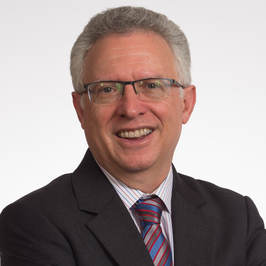 headshot of man with grey hair and glasses smiling at camera wearing gray suit and striped tie in front of white background