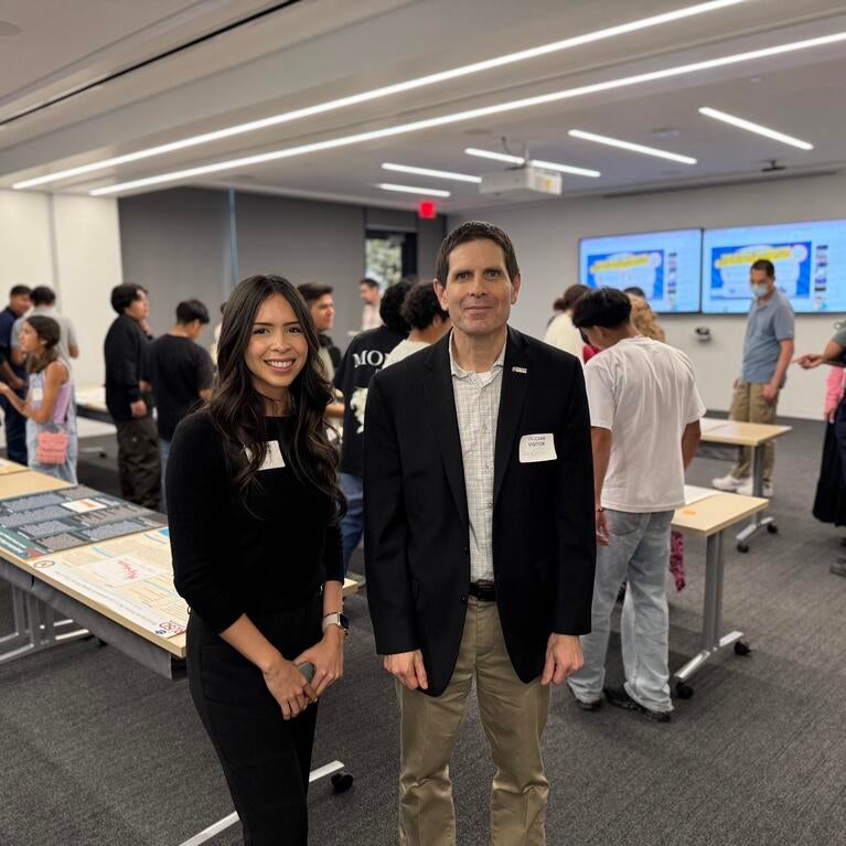Kiara Salgado, a young woman with long dark hair wearing a black outfit, stands next to Don Collins, who is wearing a black blazer and khaki pants. They are both smiling at the camera in a classroom filled with students and poster presentations displayed on tables in the background.