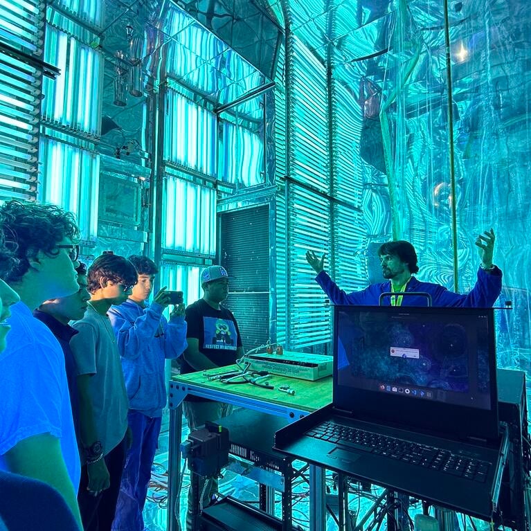 A group of five high school students on a STEPCon (Science and Technology Education Partnership Conference) tour are gathered in the world's largest indoor atmospheric processes chamber at the UC Riverside CE-CERT Atmospheric Processes Laboratory. The students are intently focused on a researcher in a blue jacket, who is enthusiastically explaining a concept with his arms raised, standing near a laptop. The massive chamber is dramatically lit by arrays of intense cyan/blue UV lights