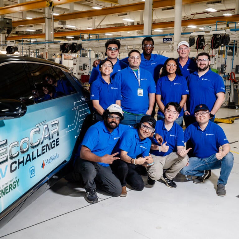 The UCR EcoCAR team gathers for a group photo next to their Cadillac LYRIQ in the workshop, all wearing matching blue team polos and smiling for the camera.