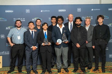 UCR EcoCAR team members stand together in formal and semi-formal attire, holding trophies in front of the official EcoCAR EV Challenge step-and-repeat backdrop featuring logos of GM, DOE, and MathWorks.