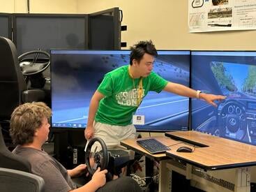A student sits in the driver's seat of the Transportation Systems Research (TSR) driving simulator, holding a steering wheel, while a researcher in a green shirt points at a large monitor displaying a driving simulation. The setup features multiple large screens surrounding the driver to create an immersive experience.
