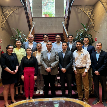 A group of 14 people stands on a staircase, posing for a photo in a well-lit room with arched windows and decorative railings.A group of 14 people stands on a staircase, posing for a photo in a well-lit room with arched windows and decorative railings.