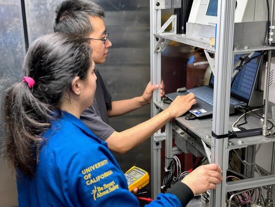 A side-view, medium shot of two UCR CE-CERT researchers working in a mobile lab truck. In the foreground, graduate student Maryam Yousefi, in a blue university jacket, holds a yellow multimeter. Behind her, graduate student Minghao Han, in a gray t-shirt and glasses, points at a laptop on a metal shelf unit filled with scientific instruments and wires.