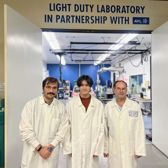Warren Norbeck stands between mentors Zisimos Toumasatos (left) and Tom Durbin (right) inside CE-CERT’s Light Duty Laboratory. All three are wearing white lab coats and are positioned under a large sign reading 'Light Duty Laboratory in Partnership with AVL.' Laboratory equipment and workstations are visible in the background.
