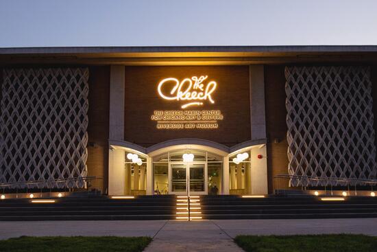 The Cheech Marin Center for Chicano Art & Culture in Riverside, California, shown at dusk with its illuminated “The Cheech” sign glowing above the entrance. Modern latticework panels flank the building’s front, and wide steps with accent lighting lead up to the glass doors.