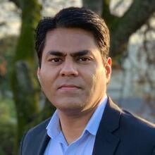 A man with short black hair, wearing a light blue collared shirt and dark blazer, looks at the camera while standing outdoors with a tree in the background.