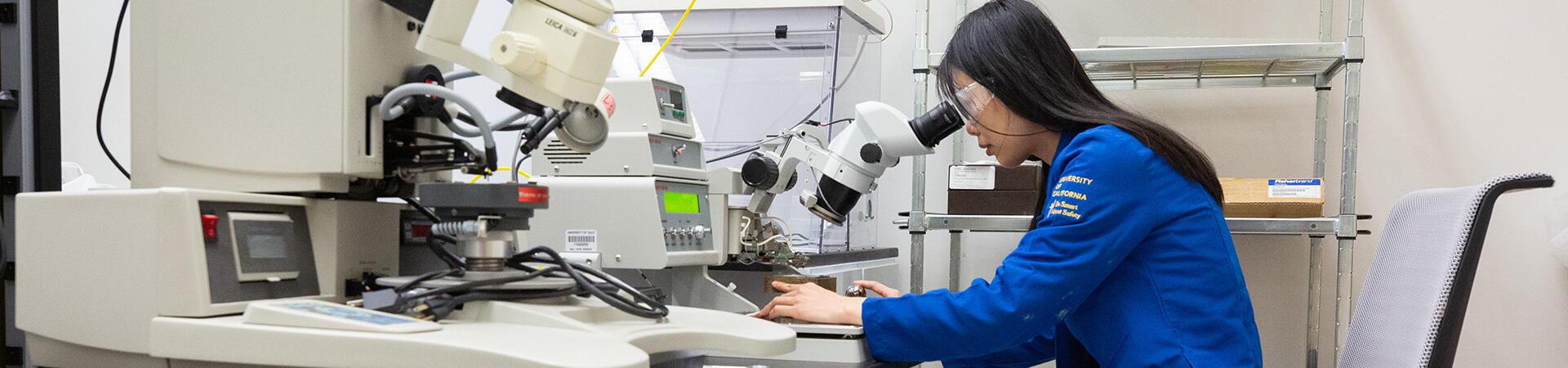 A person in a blue lab coat looks through a microscope in a research laboratory at UC Riverside.