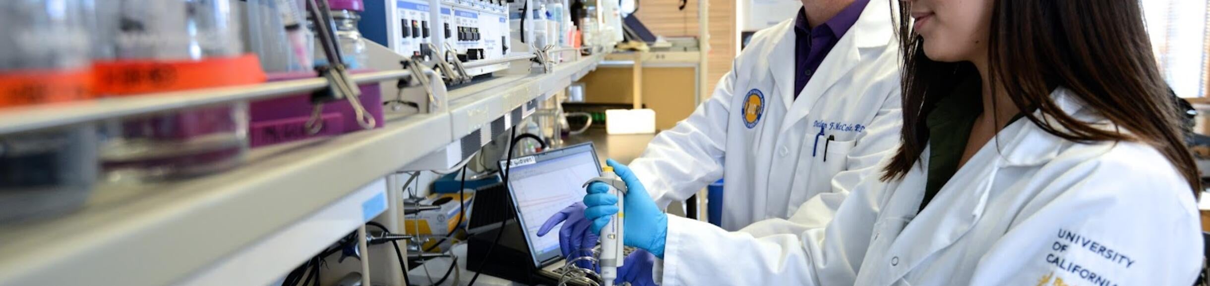 Two scientists stand in front of a row of chemicals.