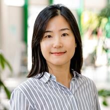 A woman with straight black hair, wearing a white button-up shirt with thin black stripes, smiles softly while standing in a bright indoor setting with plants in the background.