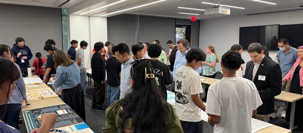 A classroom filled with students and adults viewing air quality research posters displayed on long tables. Attendees are reading, discussing, and pointing at various projects. The room is brightly lit with digital screens and projectors at the front. Don Collins is visible on the right side of the image, reviewing a student’s project.
